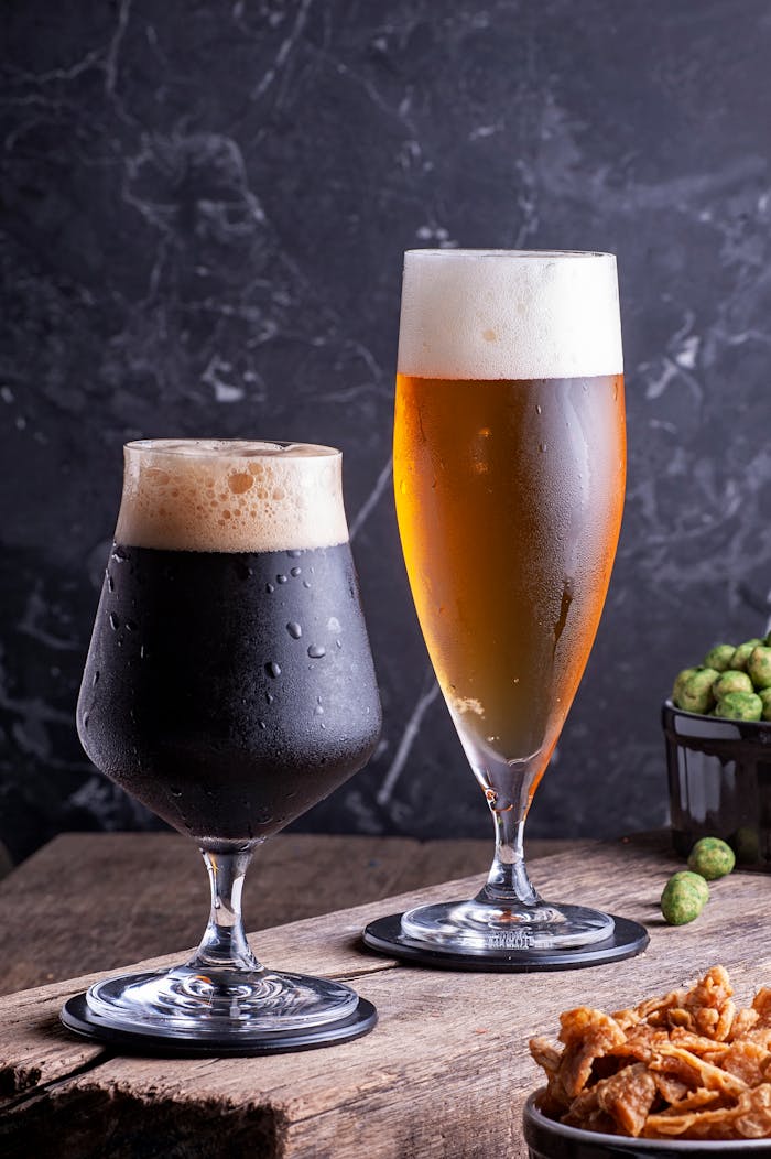 Two varieties of craft beer in glasses with snacks and hops nearby on a rustic table.